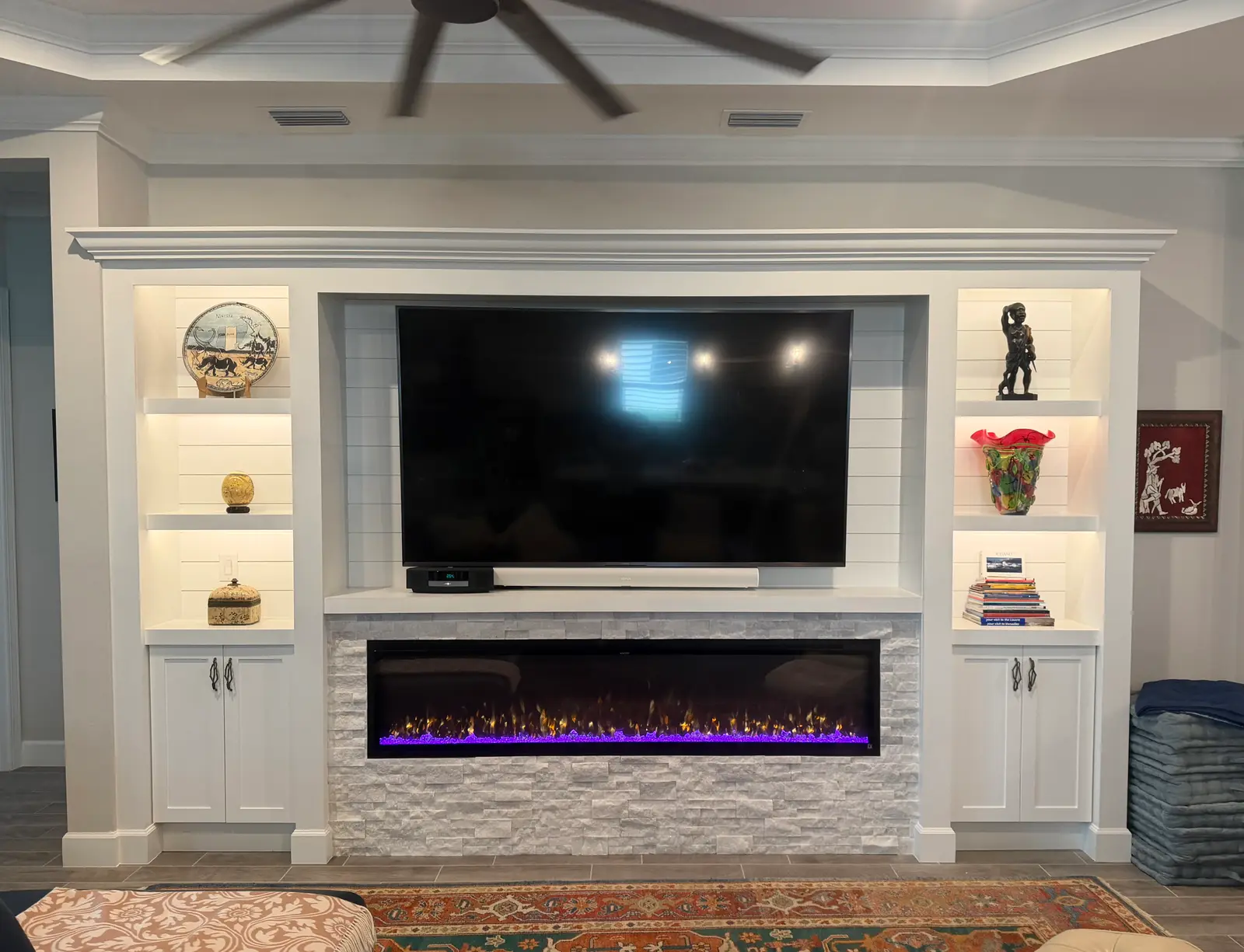 White built-in with crown molding, lit shiplap display shelves and a white stacked-stone surround around a linear electric fireplace