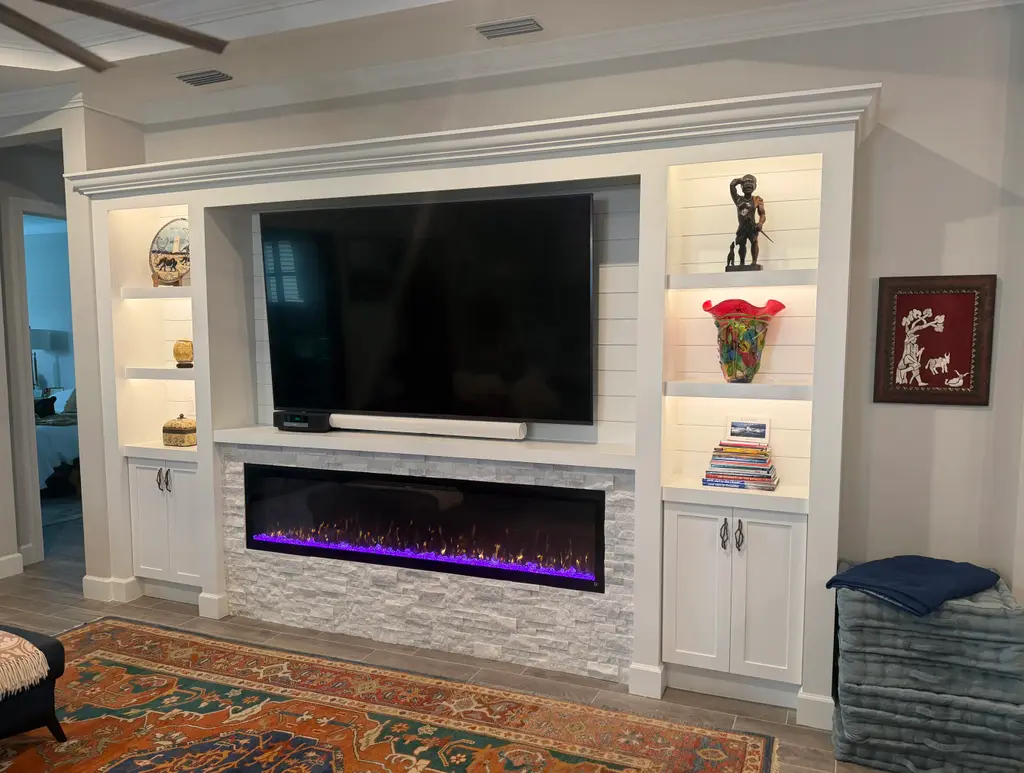 Angled view of a white crown-molded built-in with lit shelves, a wall-mounted TV and a stacked-stone linear electric fireplace