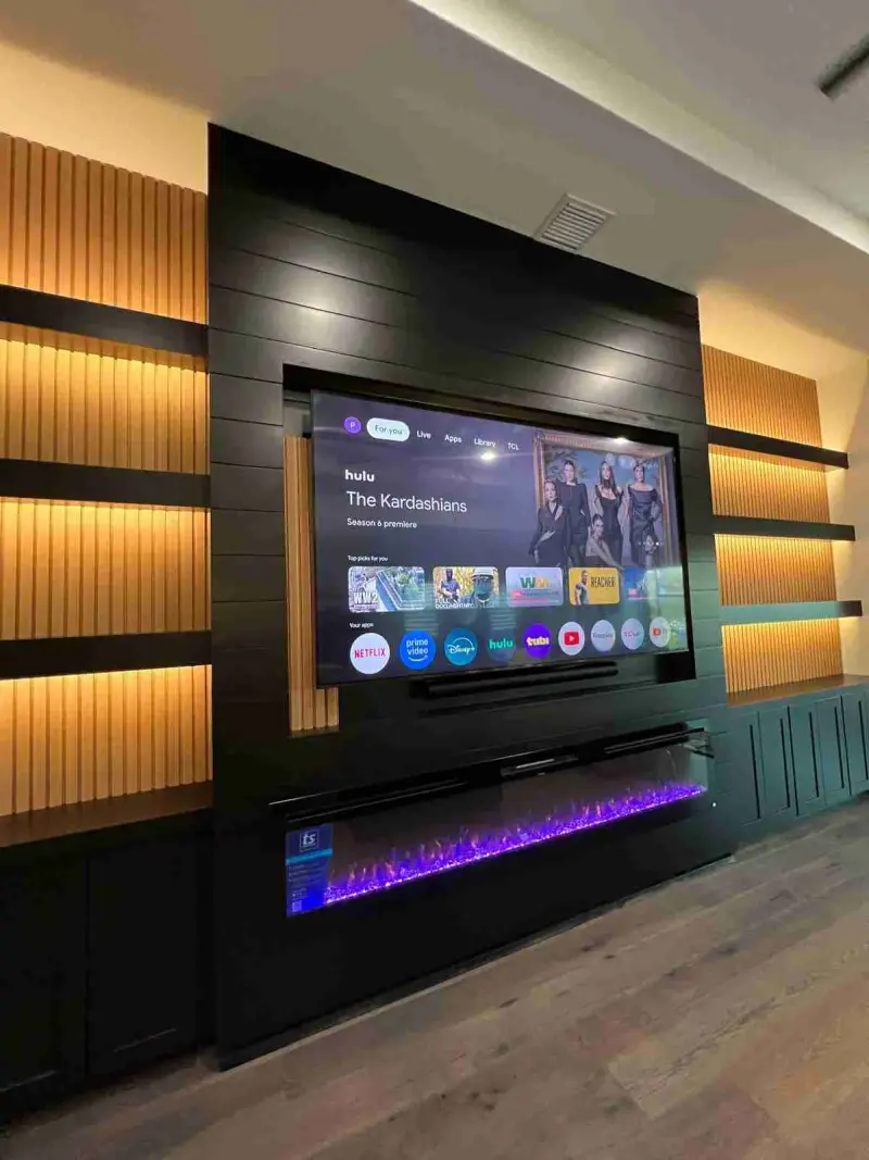 Floor-to-ceiling black shiplap media wall with backlit wood slat shelving and a long linear electric fireplace